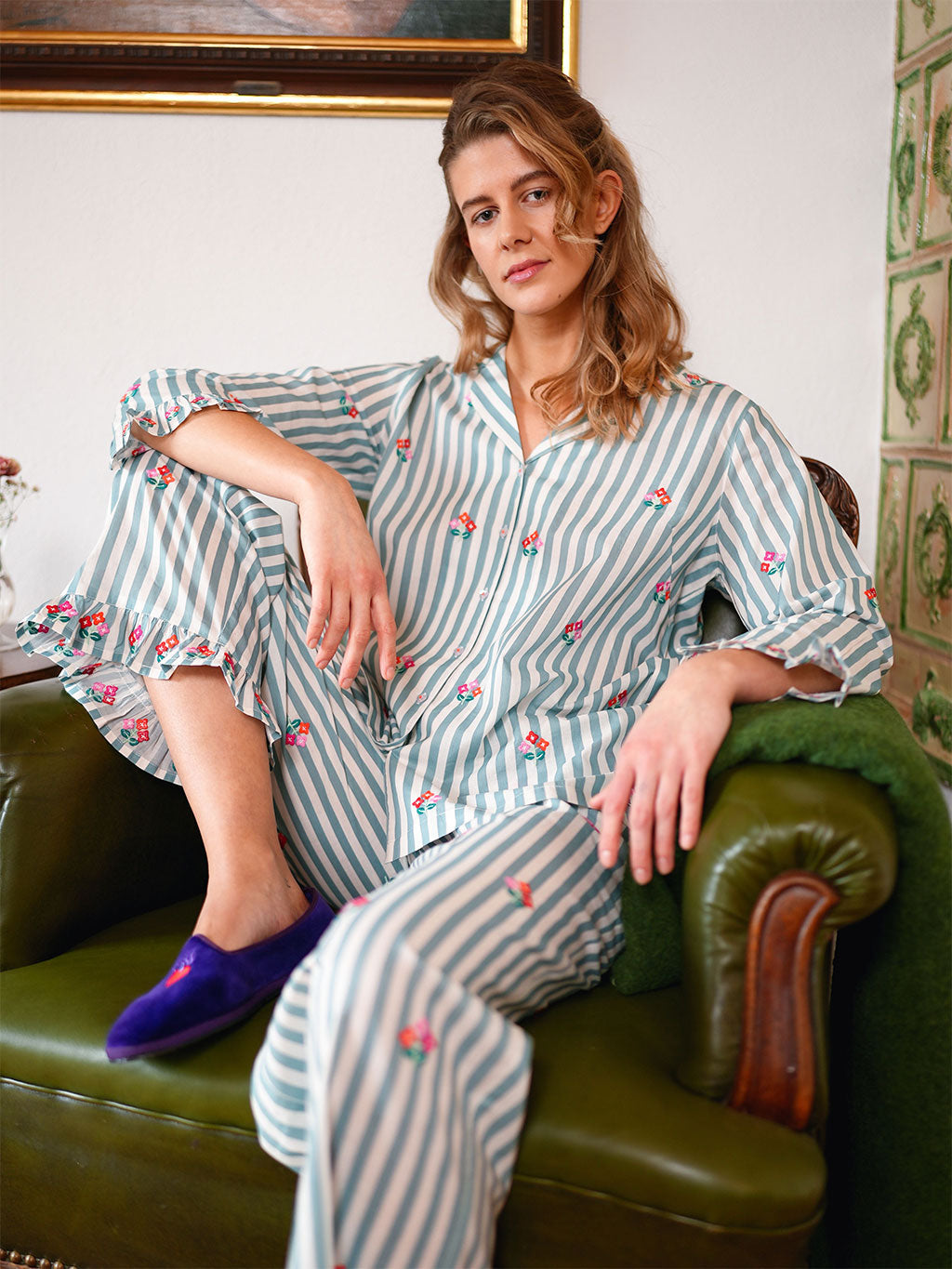 A woman sits in a luxury Nimo With Love green leather armchair wearing a matching striped pajama set with small embroidered floral details. The pajamas have vertical light blue and white stripes with scattered pink and green embroidered accents.