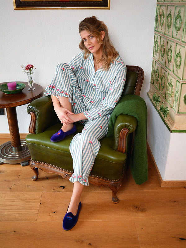 A woman sits in an olive green leather armchair wearing a luxury Nimo With Love light blue and white striped pajama set with pink floral prints and dark blue velvet slip-on shoes. The setting is a cozy room with wooden floors, a side table, and a green throw blanket draped over the chair.
