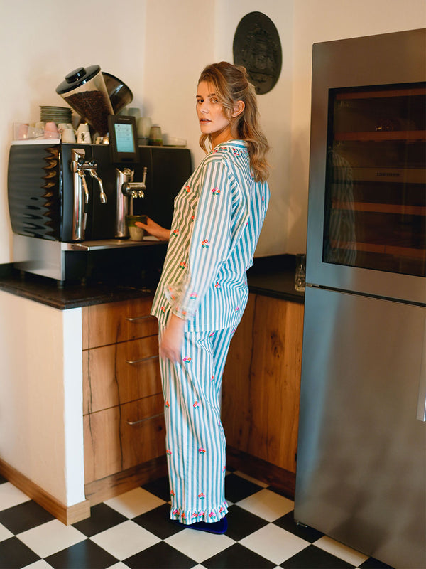 A woman wearing a luxury Nimo With Love light blue and white vertically striped pajama set with small red strawberry motifs stands in a kitchen, looking over her shoulder. The kitchen features wooden cabinets, a black coffee machine, and a stainless steel refrigerator.