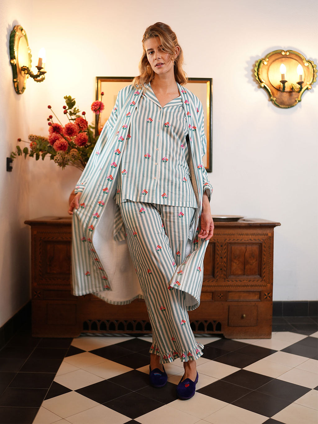 A woman models a luxury Nimo With Love matching striped pajama set with embroidered cherry details, consisting of a long-sleeve top and pants, styled with blue slip-on shoes in an indoor setting with a checkered floor and wooden furniture.