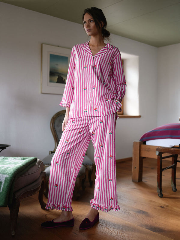 A woman stands in a bedroom wearing a luxury Nimo With Love pink and white vertically striped pajama set with small embroidered cherry motifs. The set includes a long-sleeve button-front top and matching cropped pants with ruffled hems.