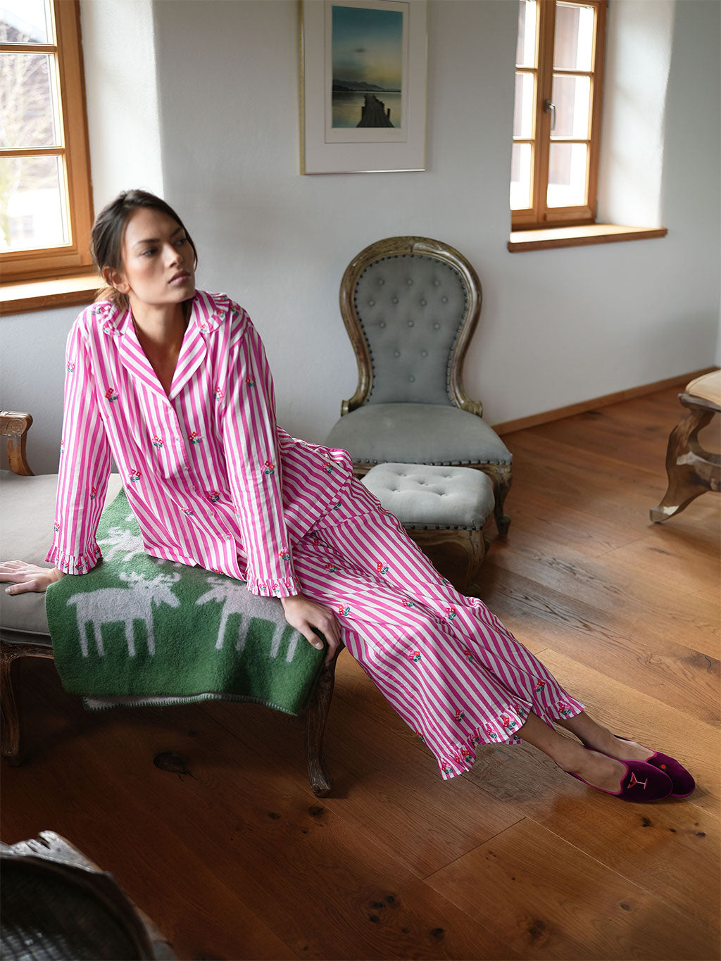 A woman wearing luxury Nimo With Love pink and white striped pajamas with a deer print sits on a green patterned throw blanket draped over a chair in a rustic room with wooden floors and antique-style furniture.