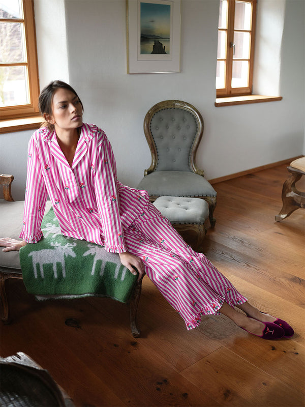 A woman wearing luxury Nimo With Love pink and white striped pajamas with a deer print sits on a green patterned throw blanket draped over a chair in a rustic room with wooden floors and antique-style furniture.