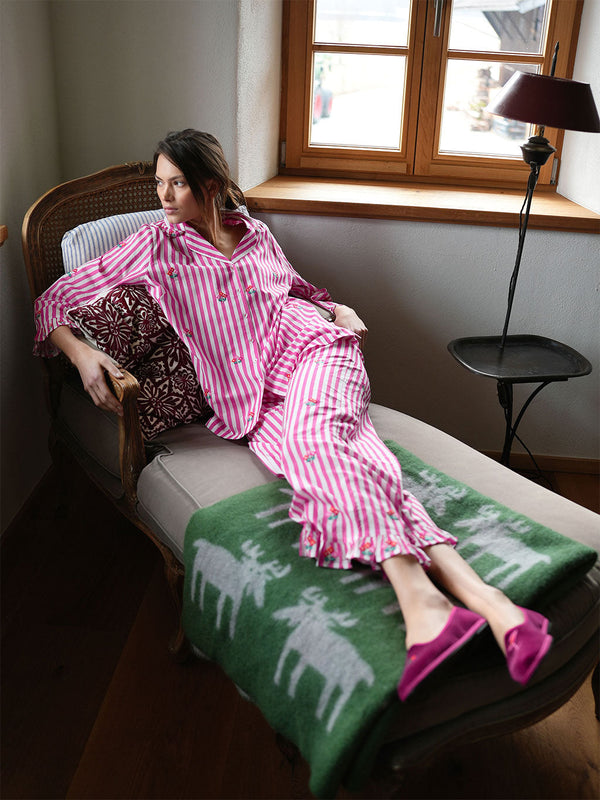 A woman reclines on a wooden daybed wearing a luxury Nimo With Love pink and white striped pajamas with small embroidered details, a green patterned blanket, and pink slip-on shoes in a softly lit room near a window.