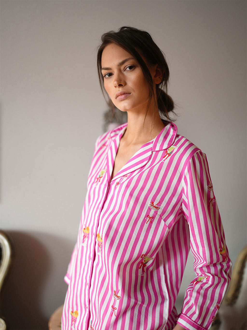 A woman wearing a luxury Nimo With Love pink and white vertically striped pajama shirt with small embroidered bow details, posed against a neutral gray wall.