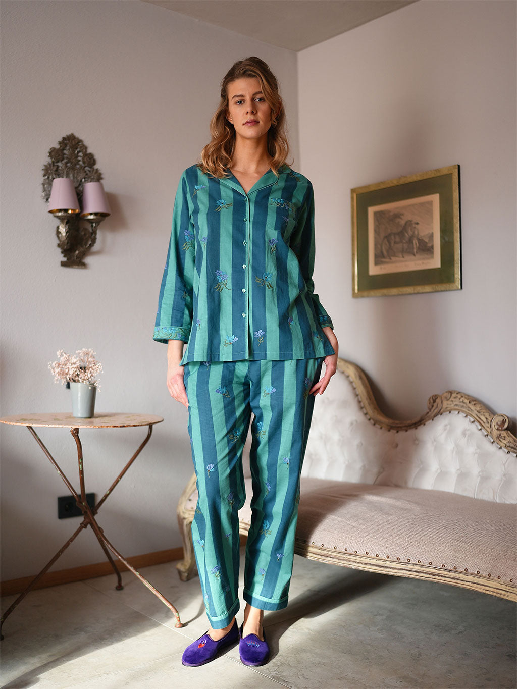 A woman modeling a luxury Nimo With Love matching two-piece pajama set with vertical teal and green stripes and small floral motifs, standing in a vintage-style room with a tufted sofa, side table, and wall decor.