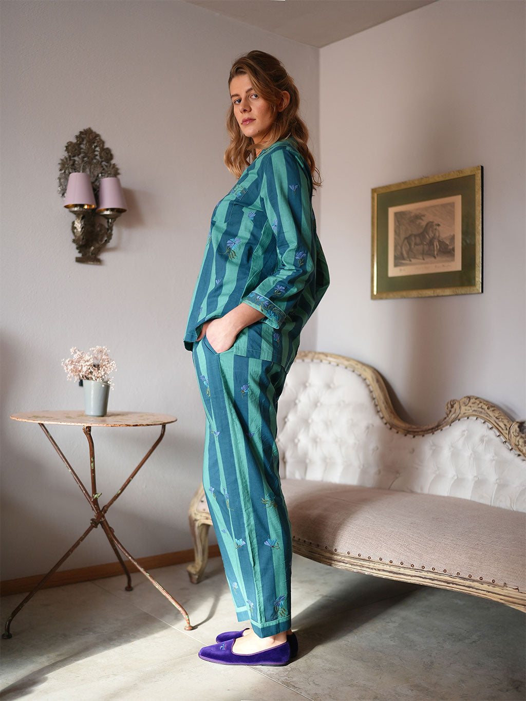 A woman wearing a luxury Nimo With Love teal and green patterned pajama set stands in a vintage-style room with a tufted sofa, side table, and framed wall art.