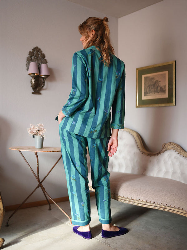 A woman stands in a room wearing a luxury Nimo With Love teal and green striped pajama set with small floral prints, paired with purple slip-on shoes. She is photographed from behind, standing next to a vintage-style chaise lounge and a small side table.