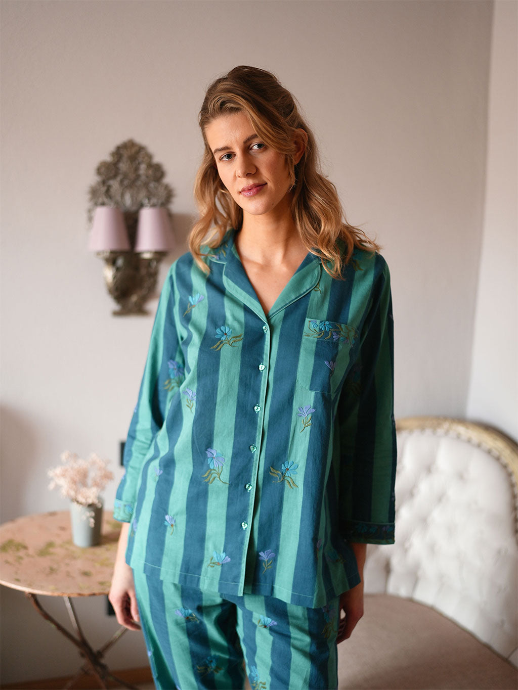 A woman modeling a luxury Nimo With Love two-piece pajama set with vertical stripes in teal and green, decorated with embroidered blue flowers and green leaves, in a softly lit bedroom setting.