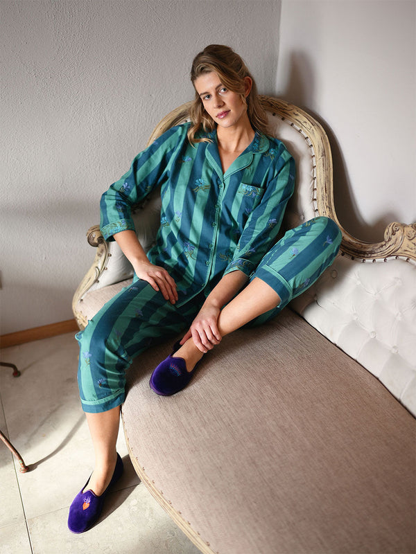 A woman sits on an upholstered chaise lounge wearing a luxury teal and green patterned Nimo With Love pajama set with purple slip-on slippers in a softly lit room.