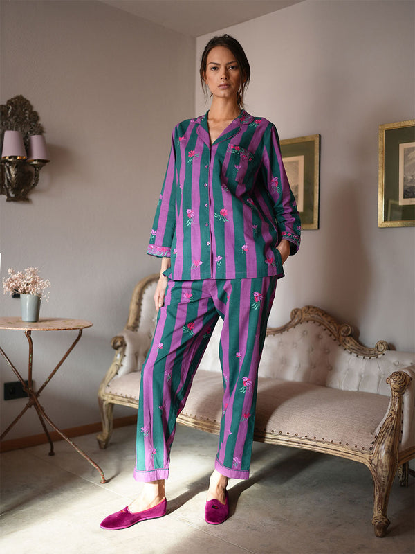 A woman models a luxury Nimo With Love matching pajama set featuring vertical stripes in teal green and magenta pink, decorated with small pink flamingos. She is wearing matching pink slip-on shoes in a vintage-style room with a chaise lounge and wall sconces.