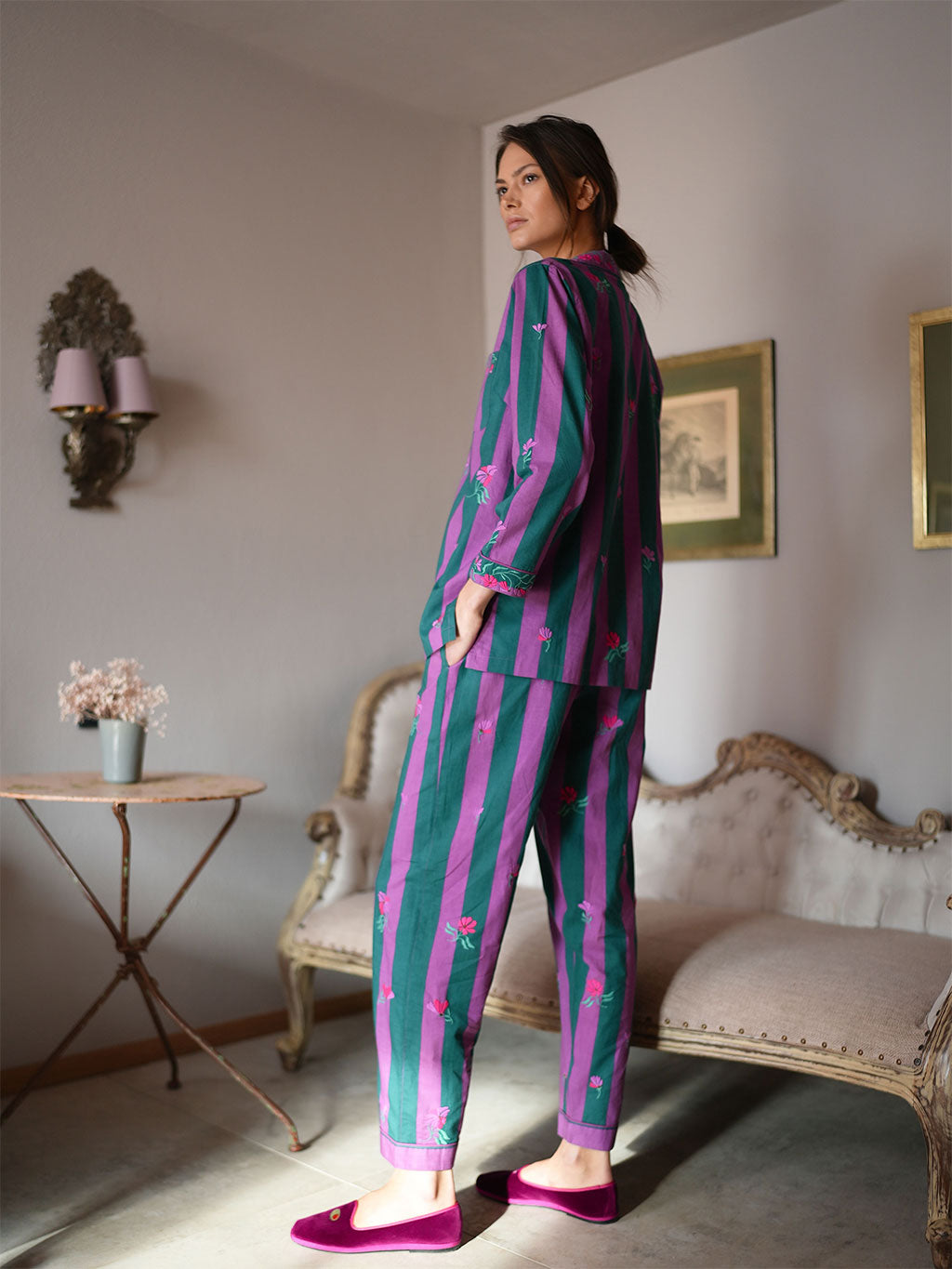 A woman stands in a vintage-style room wearing a luxury Nimo With Love matching two-piece pajama set with vertical stripes in teal green and magenta purple, decorated with small pink floral motifs. She is wearing magenta slip-on shoes.