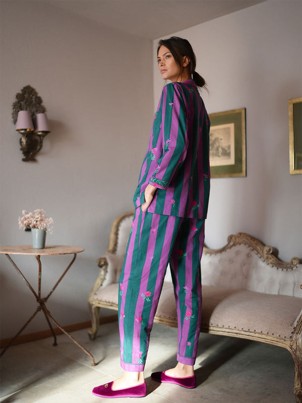 A woman stands in a vintage-style room wearing a luxury Nimo With Love matching two-piece pajama set with vertical stripes in teal green and magenta purple, decorated with small pink floral motifs. She is wearing magenta slip-on shoes.