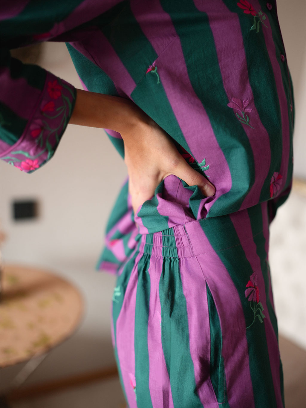 A close-up lifestyle shot of a model wearing a luxury Nimo With Love matching two-piece pajama set with vertical stripes in dark green and purple, decorated with embroidered pink flowers. The person is shown from the side, pulling the waistband of the pants.