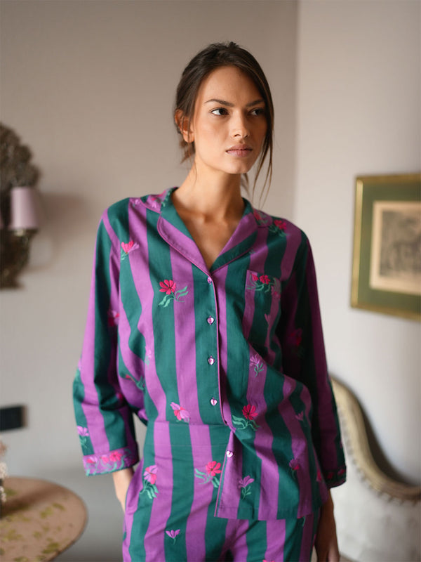 A woman wearing a luxury Nimo With Love matching pajama set with vertical purple and green stripes and pink floral embroidery, standing in a softly lit indoor room.