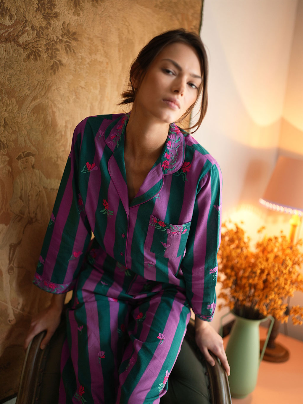 A woman wearing a luxury Nimo With Love purple and green striped GOTS certified cotton pajama set with pink floral embroidery, posed in a warmly lit room with a patterned wallpaper background and a lamp.