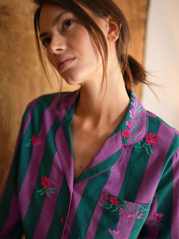 A close-up of a woman wearing a luxury Nimo With Love purple and green striped button-up pajama shirt with pink floral embroidery on the collar and chest pocket.