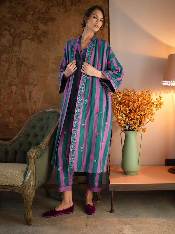 A woman models a luxury Nimo With Love matching two-piece kurta set with vertical stripes in purple, teal, and green, featuring floral embroidery down the front. She is wearing matching striped trousers and dark purple velvet slip-on shoes in a styled room setting with a lamp, a vase of yellow flowers, and an armchair.