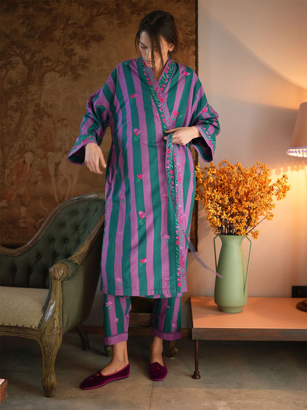 A woman wearing a luxury Nimo With Love matching two-piece pajama set with vertical pink and teal stripes and floral embroidery, standing in a warmly lit room next to a green armchair and a vase of yellow flowers.