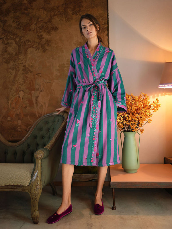 A woman models a luxury Nimo With Love long-sleeved striped bathrobe with pink and teal vertical stripes and embroidered details, paired with dark plum velvet slip-on shoes in a vintage-style room setting.