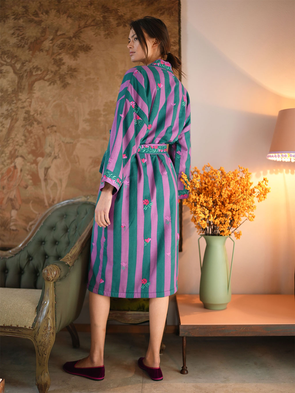 A woman wearing a luxury Nimo With Love long-sleeved striped bathrobe with pink and teal vertical stripes, standing in a warmly lit room next to a green tufted armchair and a vase of yellow flowers.