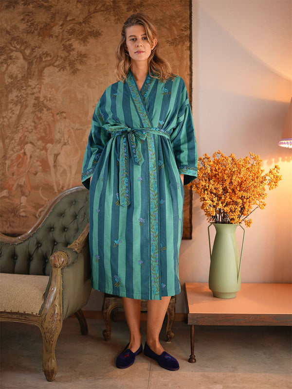 A woman wearing a teal and green vertically striped silk-like robe with embroidered details by Nimo With Love, standing in a vintage room next to a green tufted armchair and a table with a vase of orange flowers.