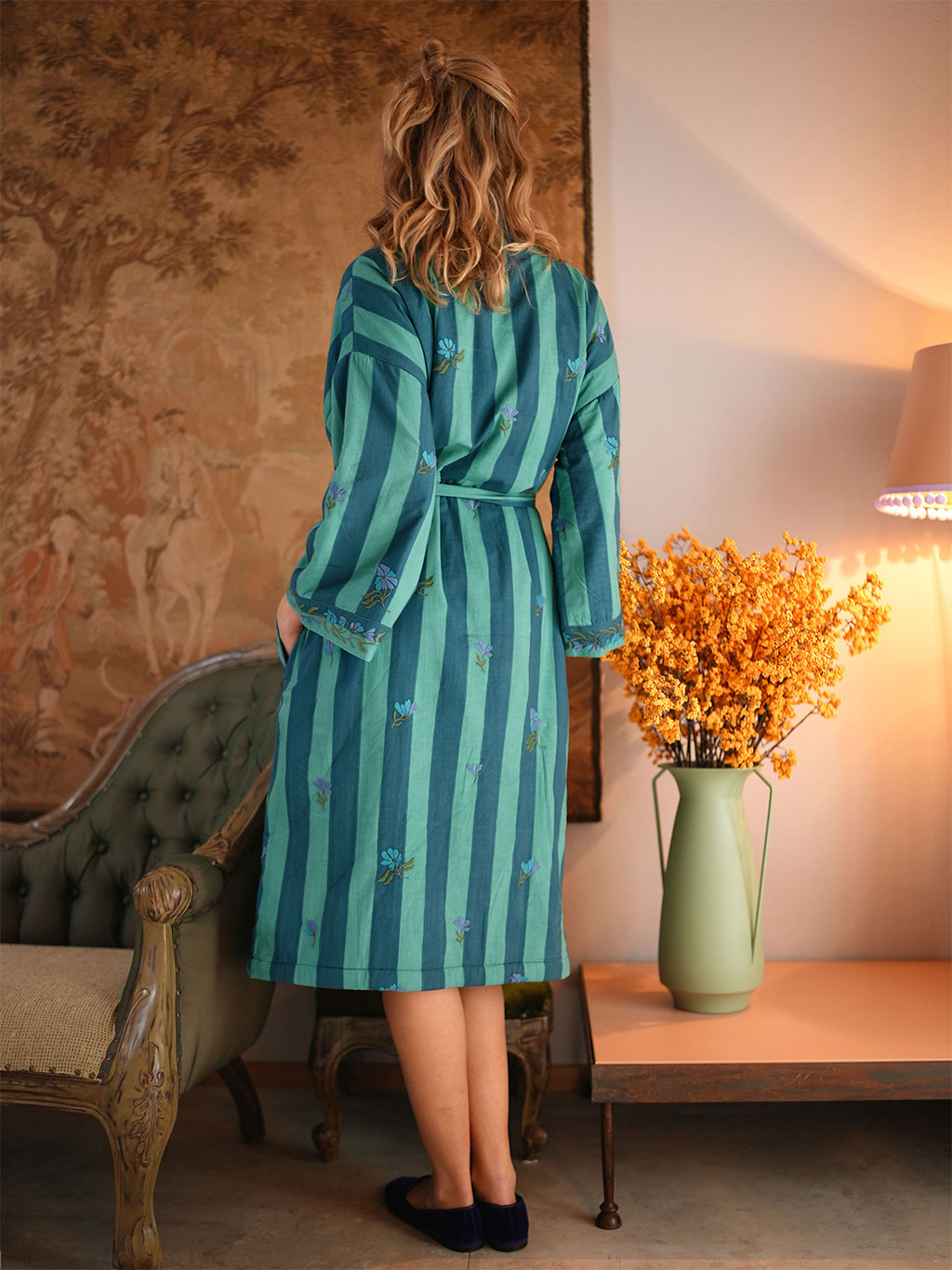 A woman stands with her back to the camera wearing a Nimo With Love teal and green vertically striped robe with embroidered blue floral details. She is in a warmly lit room with a vintage green armchair, a side table with a vase of yellow flowers, and a wall tapestry.