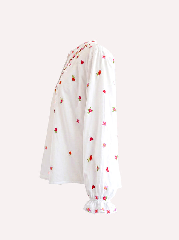 White Nimo With Love long-sleeve blouse with small red and green strawberry embroidery, featuring a gathered ruffled hem and cuffs, shown from a side profile on a light beige background.