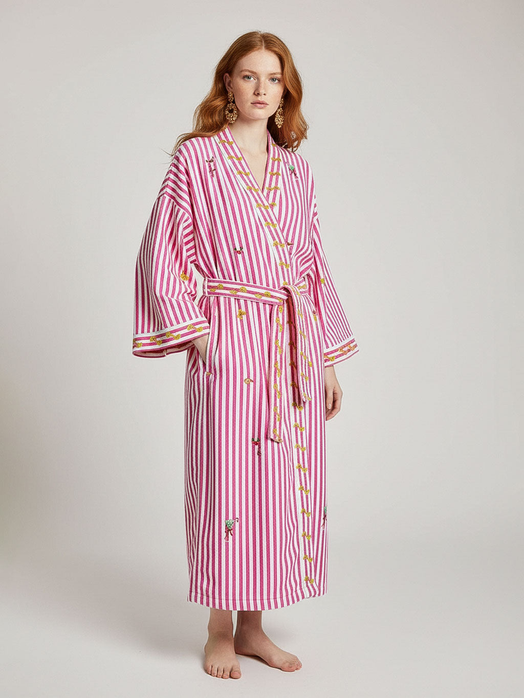 A woman modeling a luxury Nimo With Love pink and white vertically striped bathrobe with embroidered details, standing against a light gray studio background.