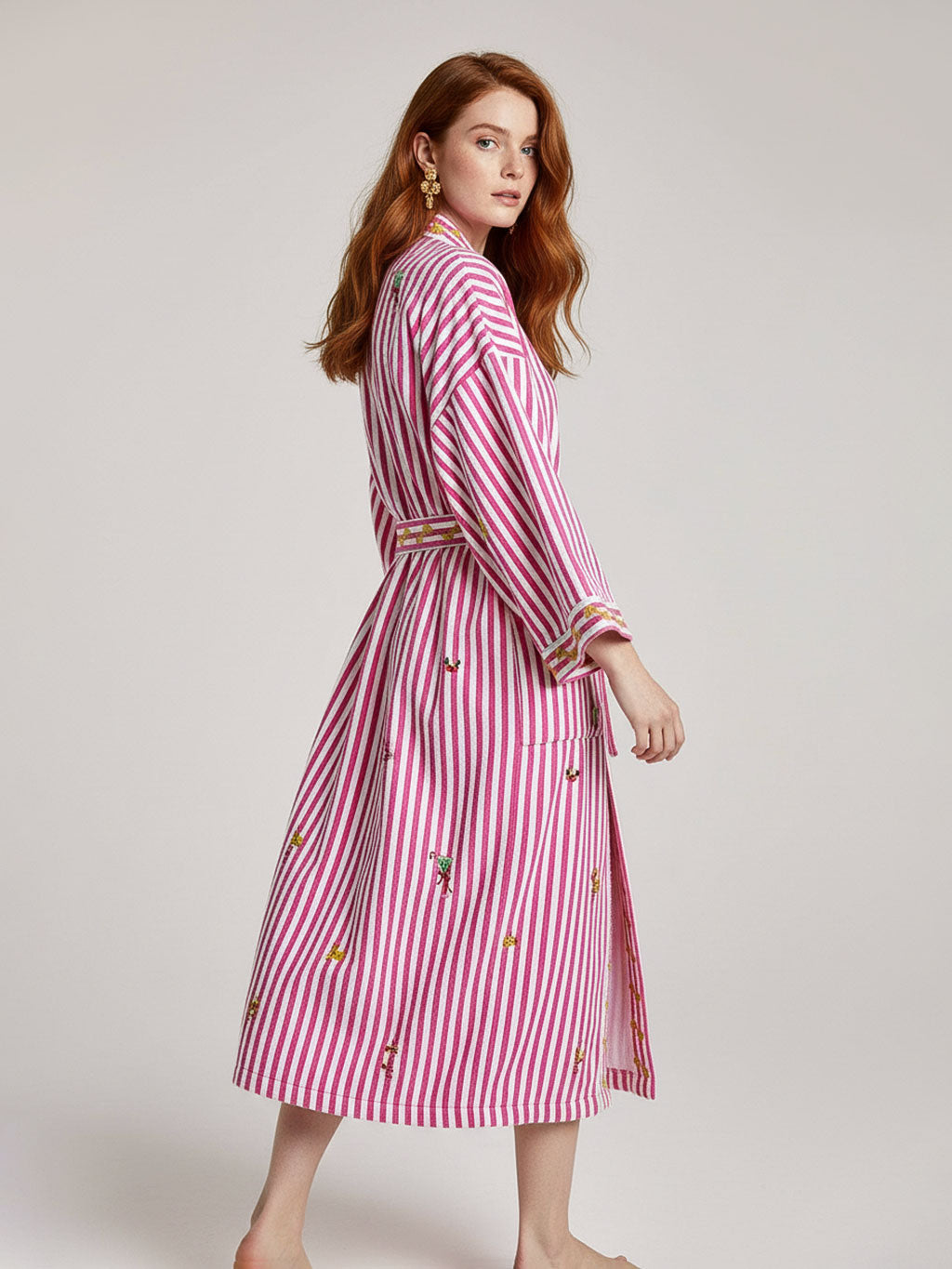 A woman modeling a luxury Nimo With Love pink and white vertically striped midi dress with a matching belt and small embroidered details, photographed against a plain light gray studio background.