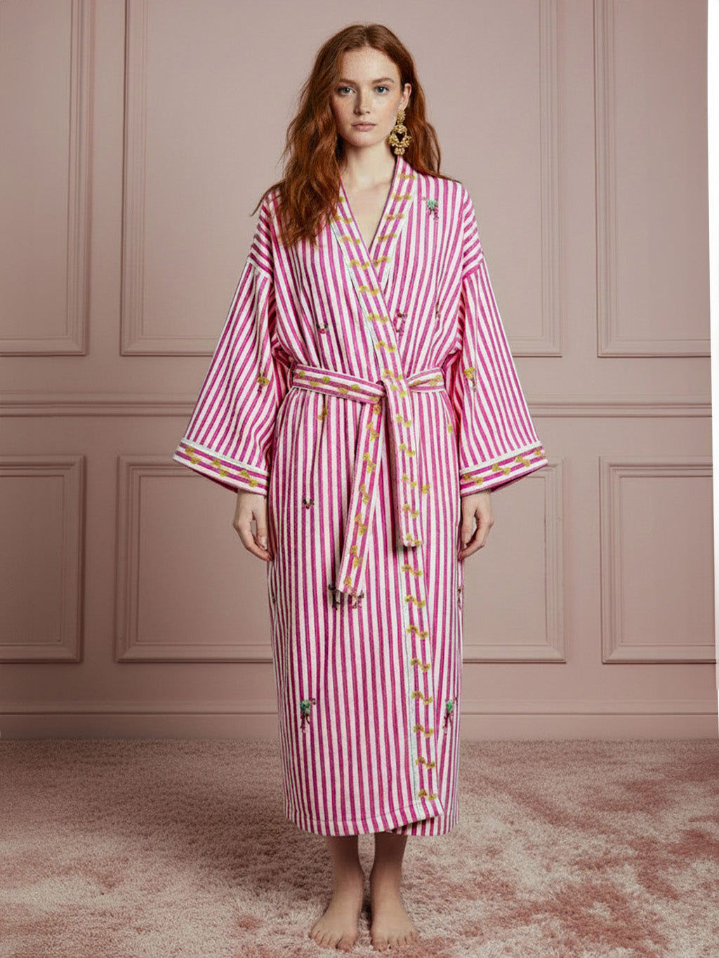 A woman stands barefoot in a luxury Nimo With Love pink robe with vertical pink and white stripes, gold trim, and embroidered floral details, posed against a pink paneled wall and textured pink rug.
