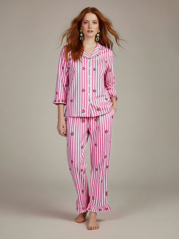A woman modeling a luxury Nimo With Love pink and white striped pajama set with small cherry prints, standing against a neutral beige studio background.