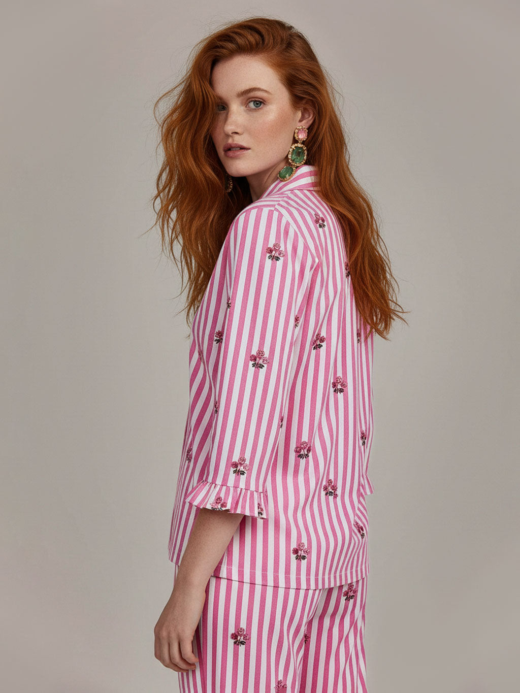 A woman with long wavy red hair is shown from the side/back wearing a luxury Nimo With Love pink and white striped shirt and matching striped pants with small embroidered floral details. She is wearing large green earrings against a plain light gray studio background.