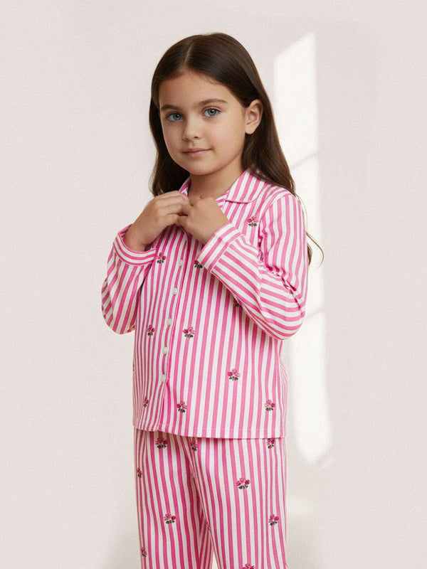 A young girl with long brown hair wearing a luxury Nimo With Love pink and white vertically striped pajama set with small embroidered floral details, posing against a light beige background.
