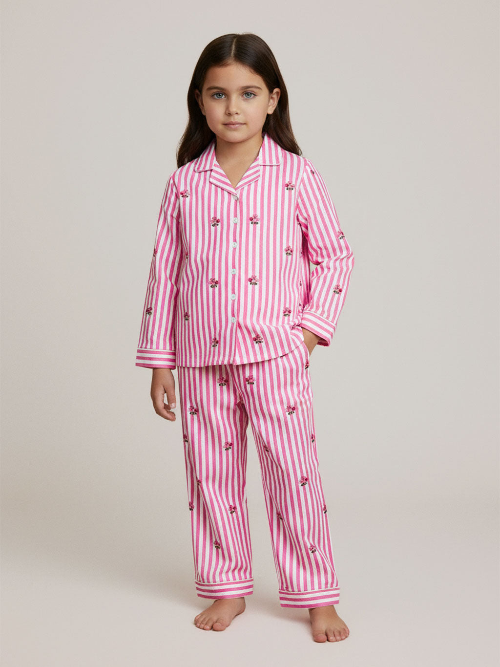 A young girl stands barefoot against a plain light beige studio background, wearing a luxury Nimo With Love pink and white vertically striped pajama set with small embroidered floral details. The set includes a long-sleeve button-front top and matching pants.