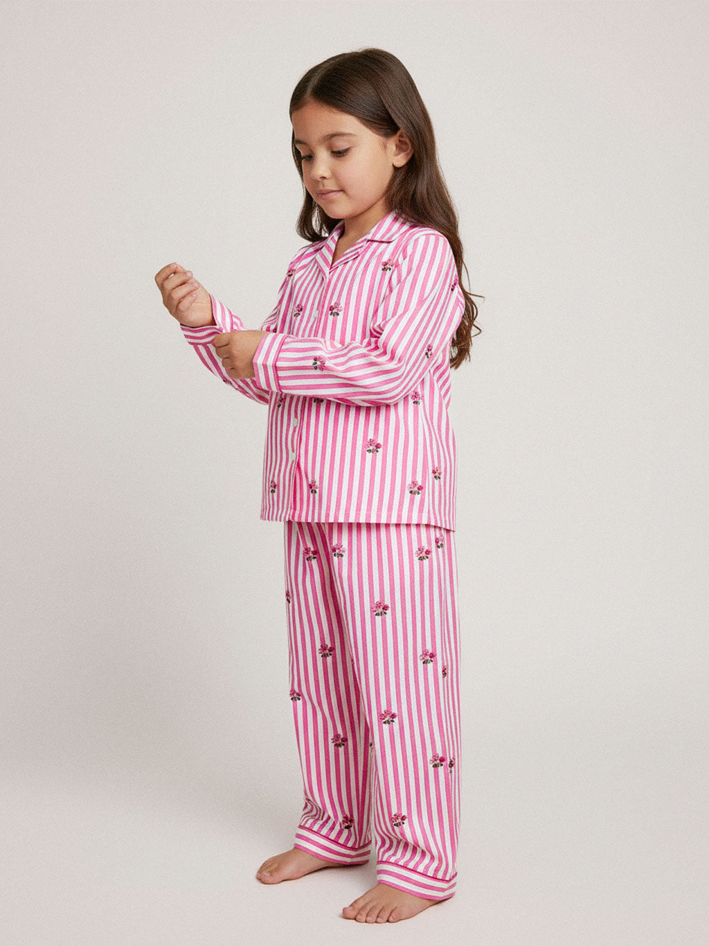 A young girl with long brown hair stands barefoot against a light gray studio background, wearing a luxury Nimo With Love pink and white vertically striped pajama set with small embroidered cherry motifs.