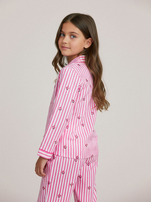 A young girl with long brown hair is shown from the back wearing a luxury Nimo With Love pink and white striped pajama set with embroidered cherries. The background is a solid light gray studio backdrop.