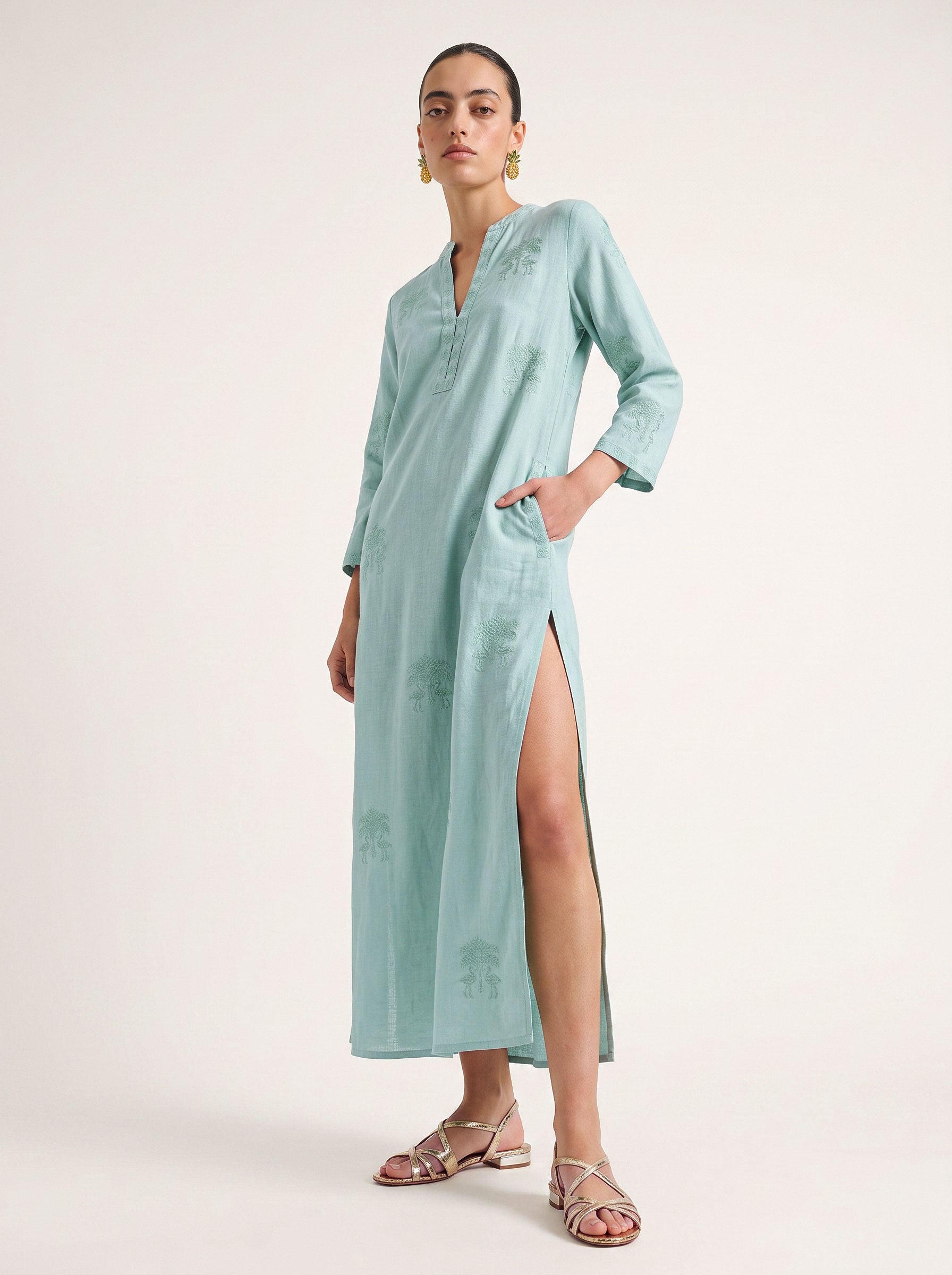 A woman modeling a light mint green maxi kurta maxi dress with a high side slit, long sleeves, and subtle tonal embroidery, paired with metallic sandals against a plain off-white studio background.