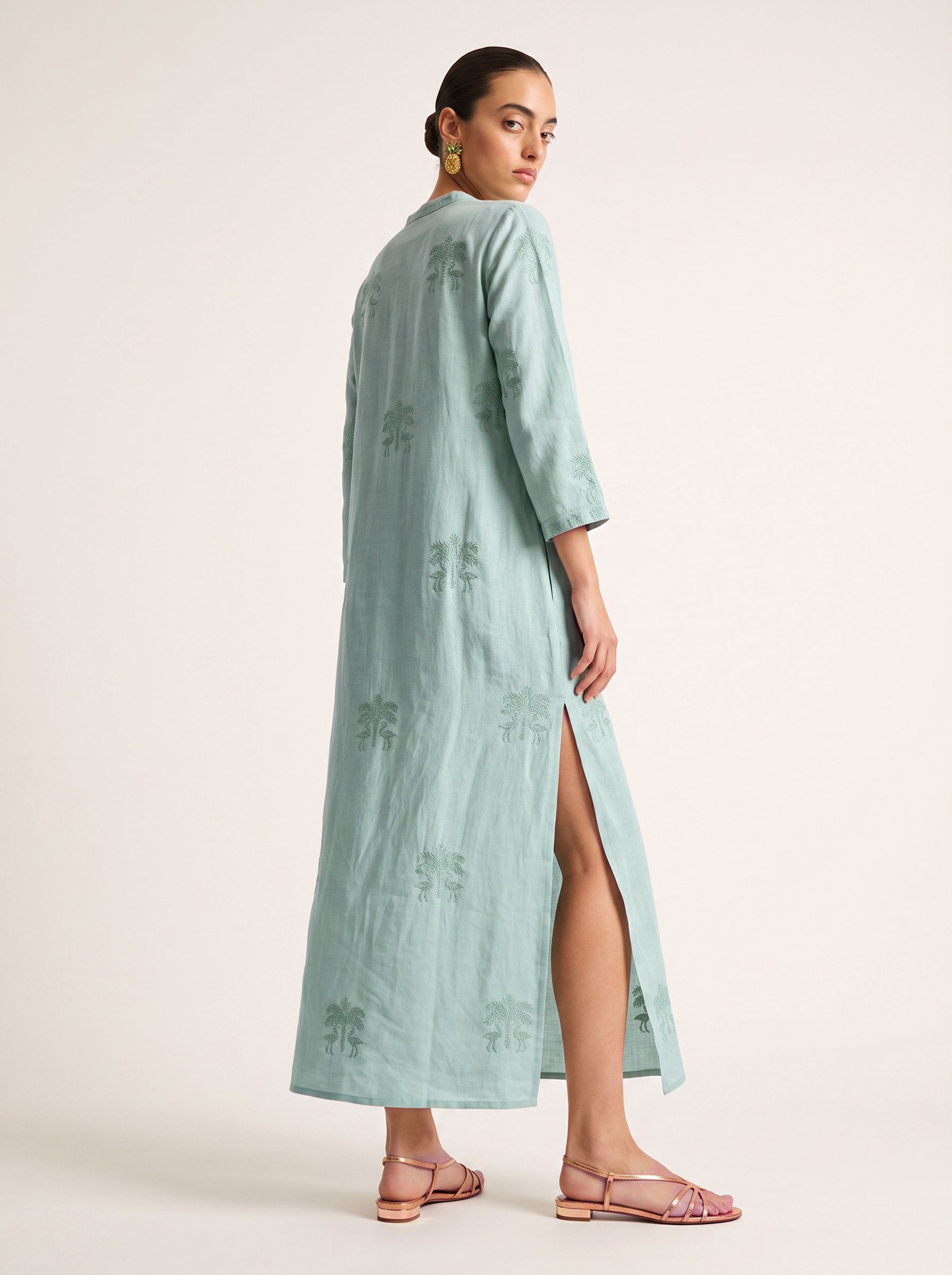 A woman modeling a long, mint green kaftan with a high side slit, shown from a three-quarter back angle against a plain light beige studio background.